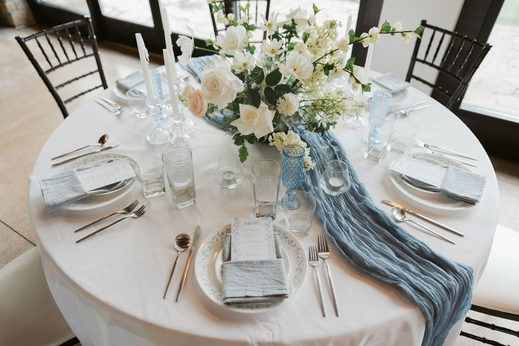 Elegant table setting with white flowers, blue runner, and silverware on a round table.